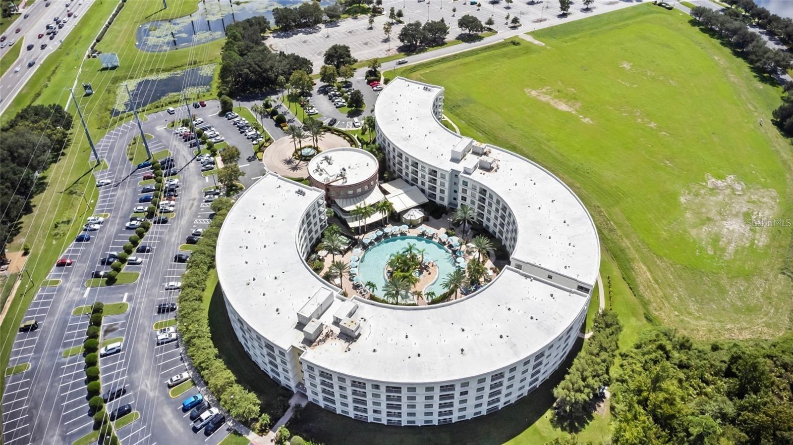 Curved multi-story building surrounding a pool, with parking lots and sports fields nearby.