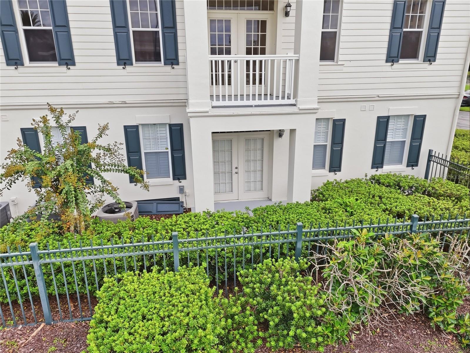 White-sided residence with blue shutters, an entry balcony, and hedges behind a metal fence.