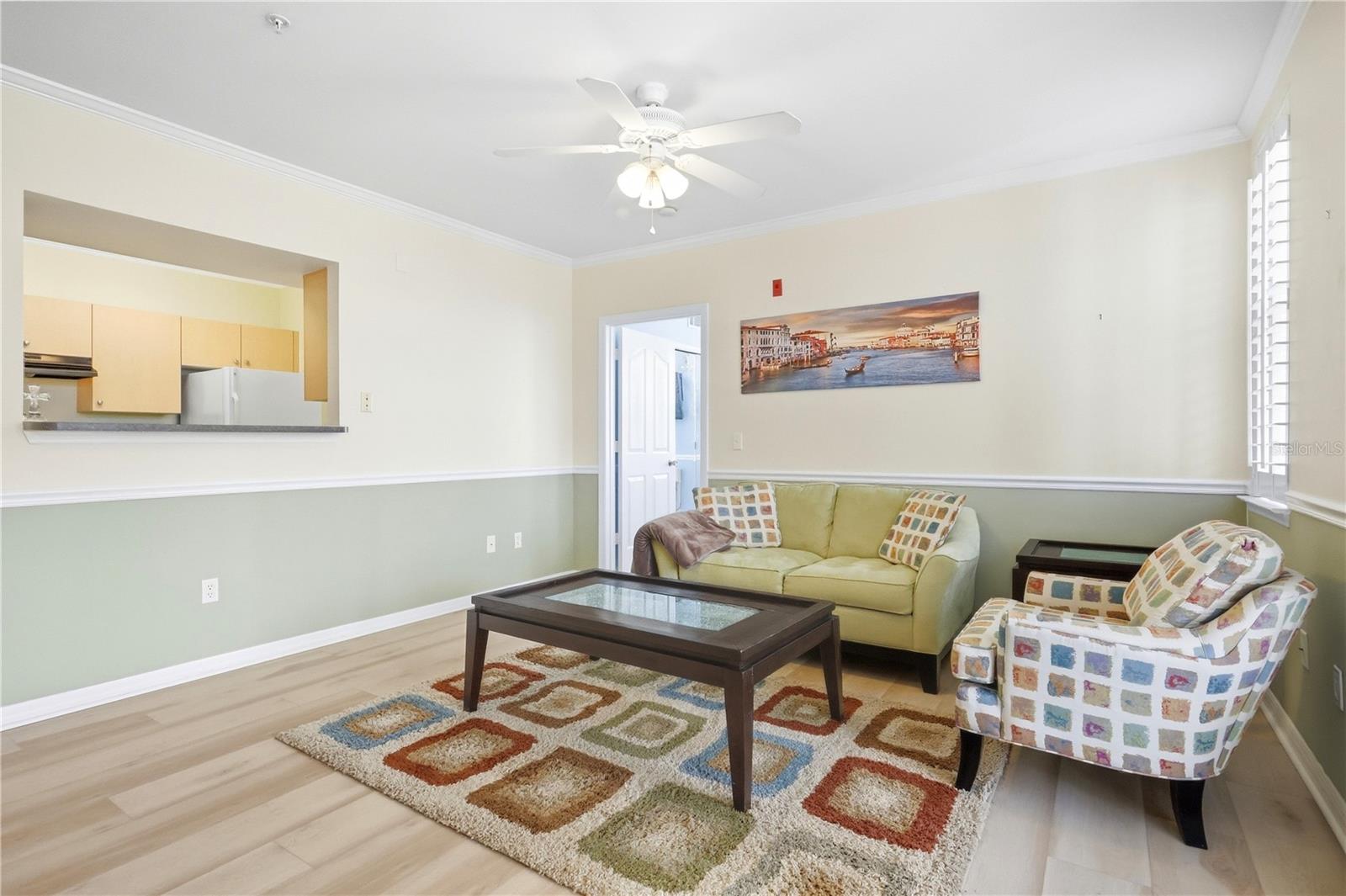 Living room with pale green walls, patterned seating, a glass-top coffee table, and a kitchen pass-through.