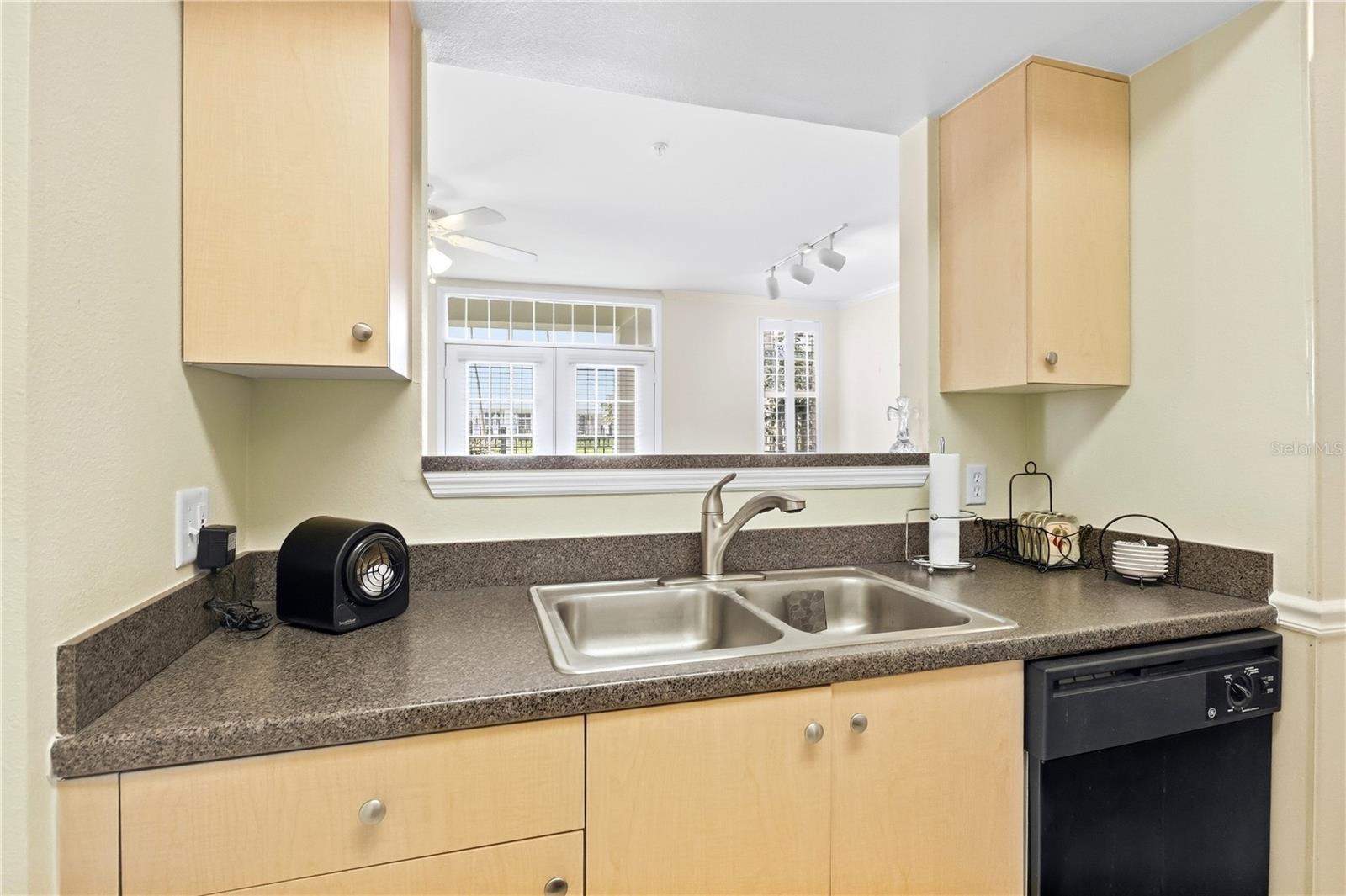 Kitchen with light wood cabinets, speckled counters, a double-basin sink, and a windowed pass-through.