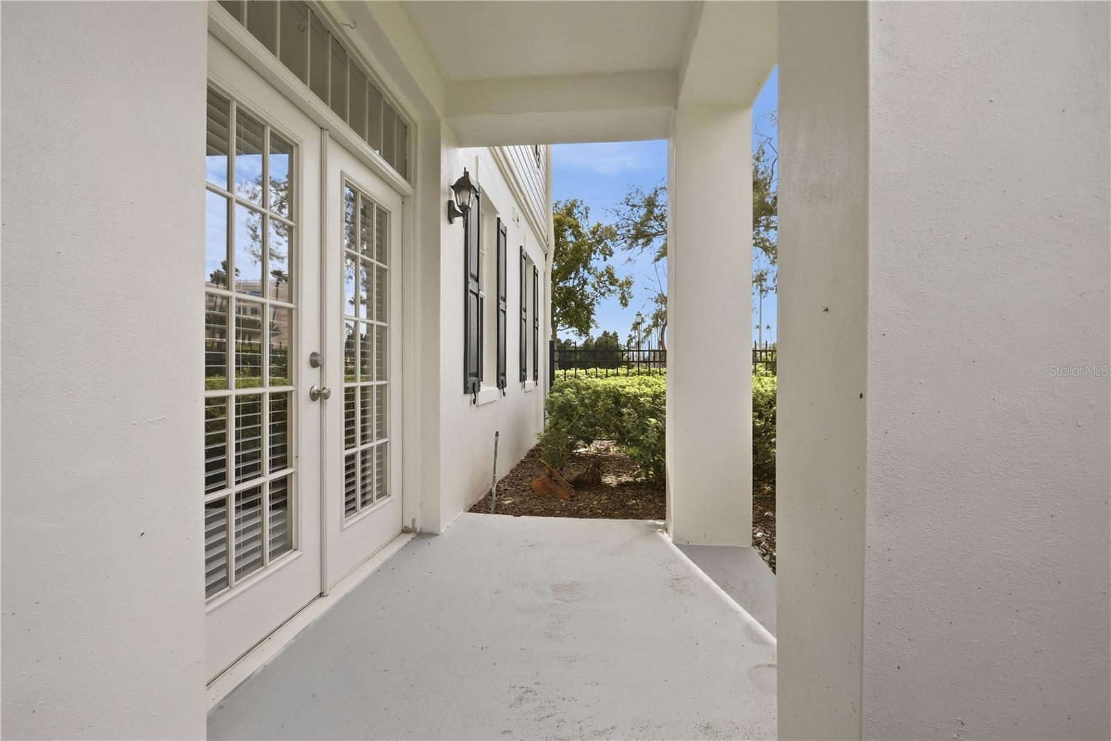 Covered concrete patio beside white French doors, with shutters and a hedge-lined yard.