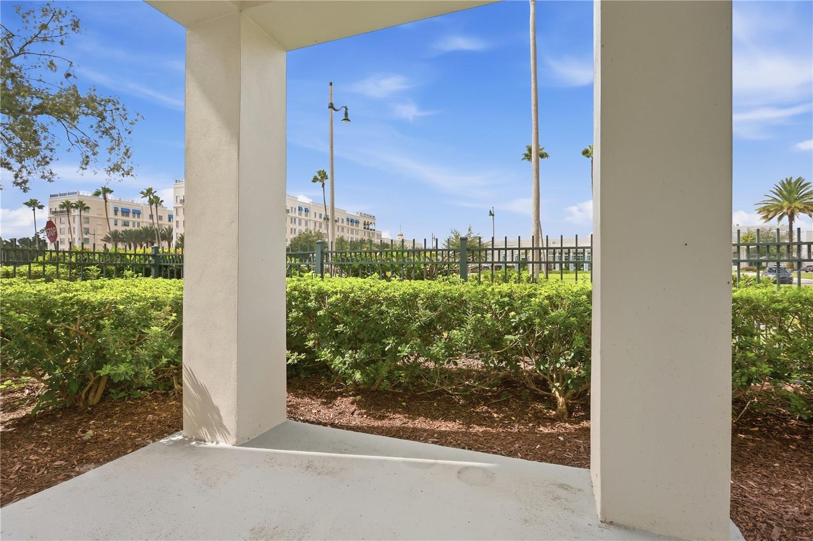 Covered patio with stucco columns overlooking hedges, a metal fence, and palm trees.