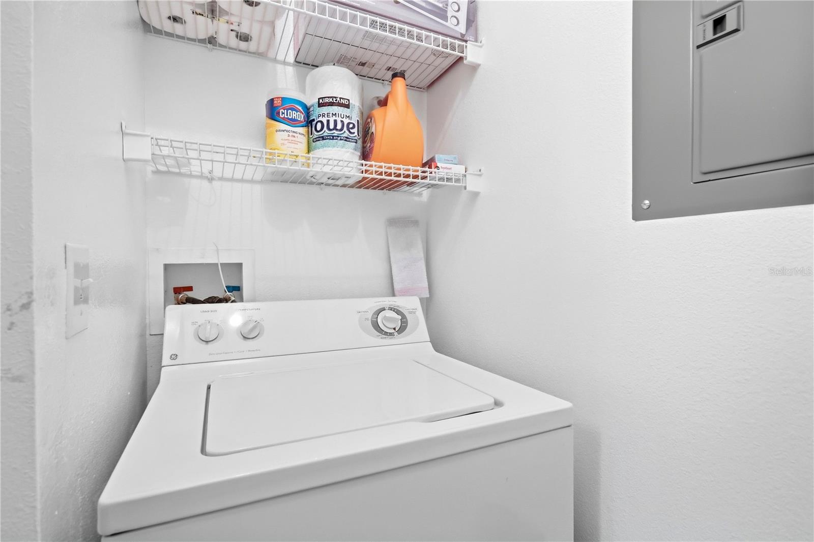 Laundry room with a top-loading washer, stocked wire shelves, and a wall-mounted electrical panel.