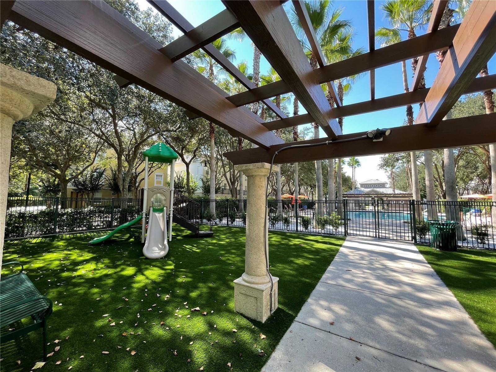 Community courtyard with a pergola and playground beside a fenced swimming pool.
