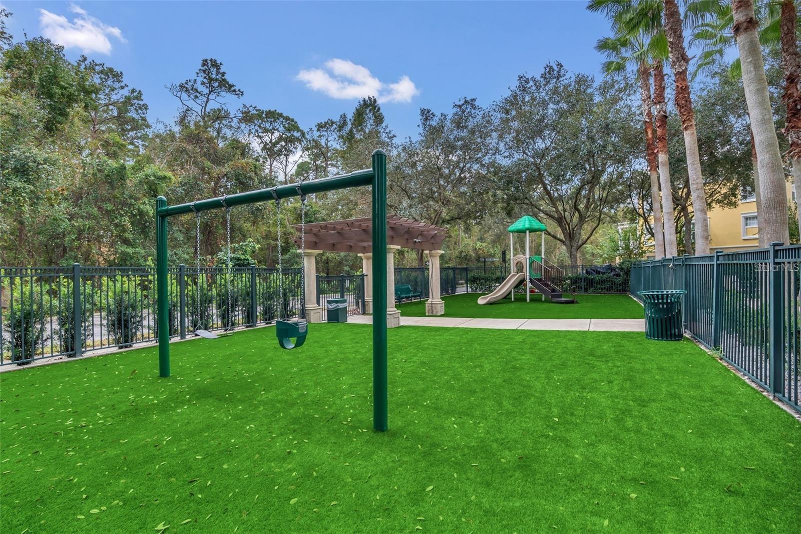 Community playground with swings and a small slide on green turf beneath mature trees.