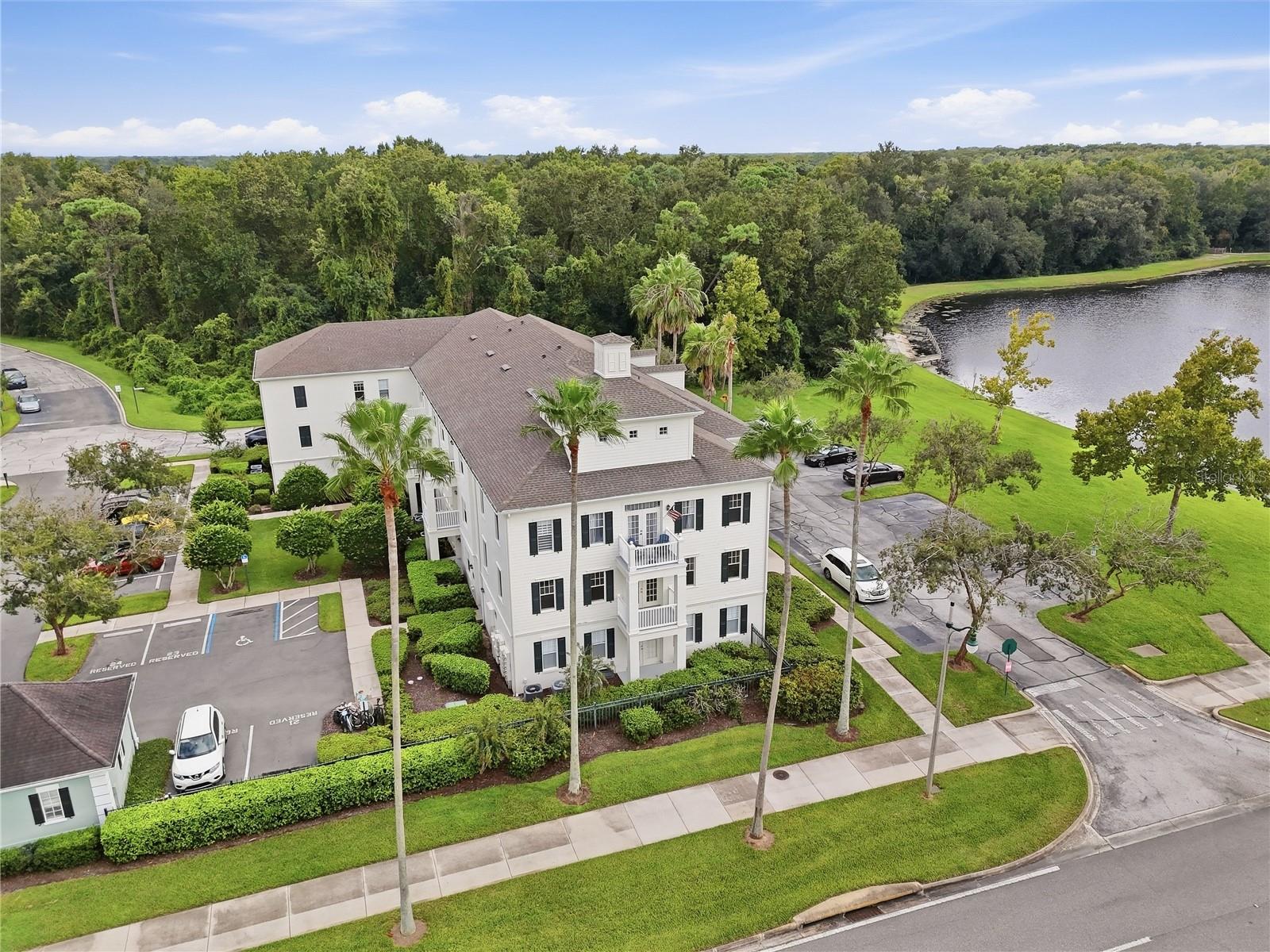 Aerial view of a white multi-story building beside a lake, with balconies and palm trees.