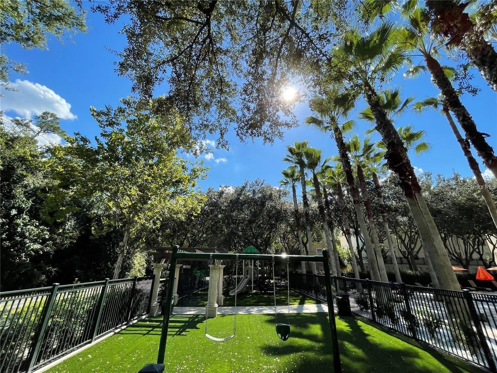 Community playground with swings on green turf beneath palm trees.