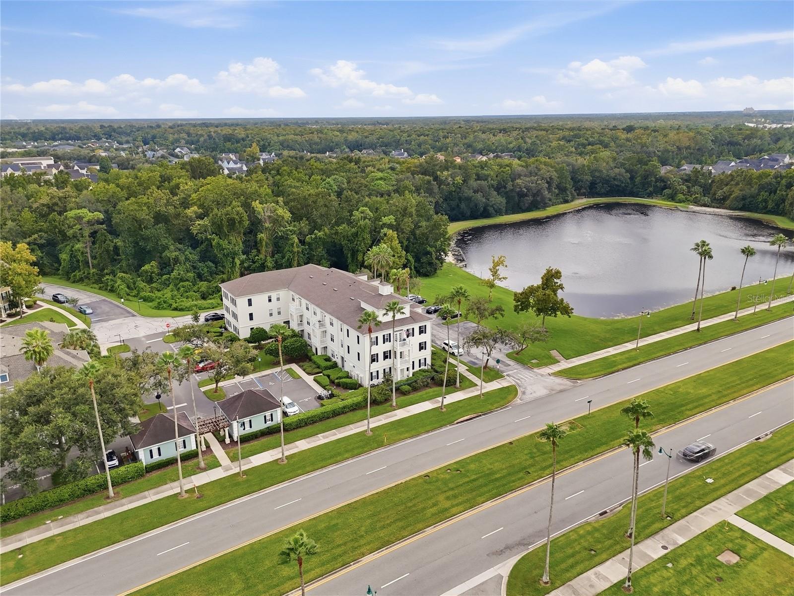 Aerial view of a residential building beside a pond and palm-lined roads.