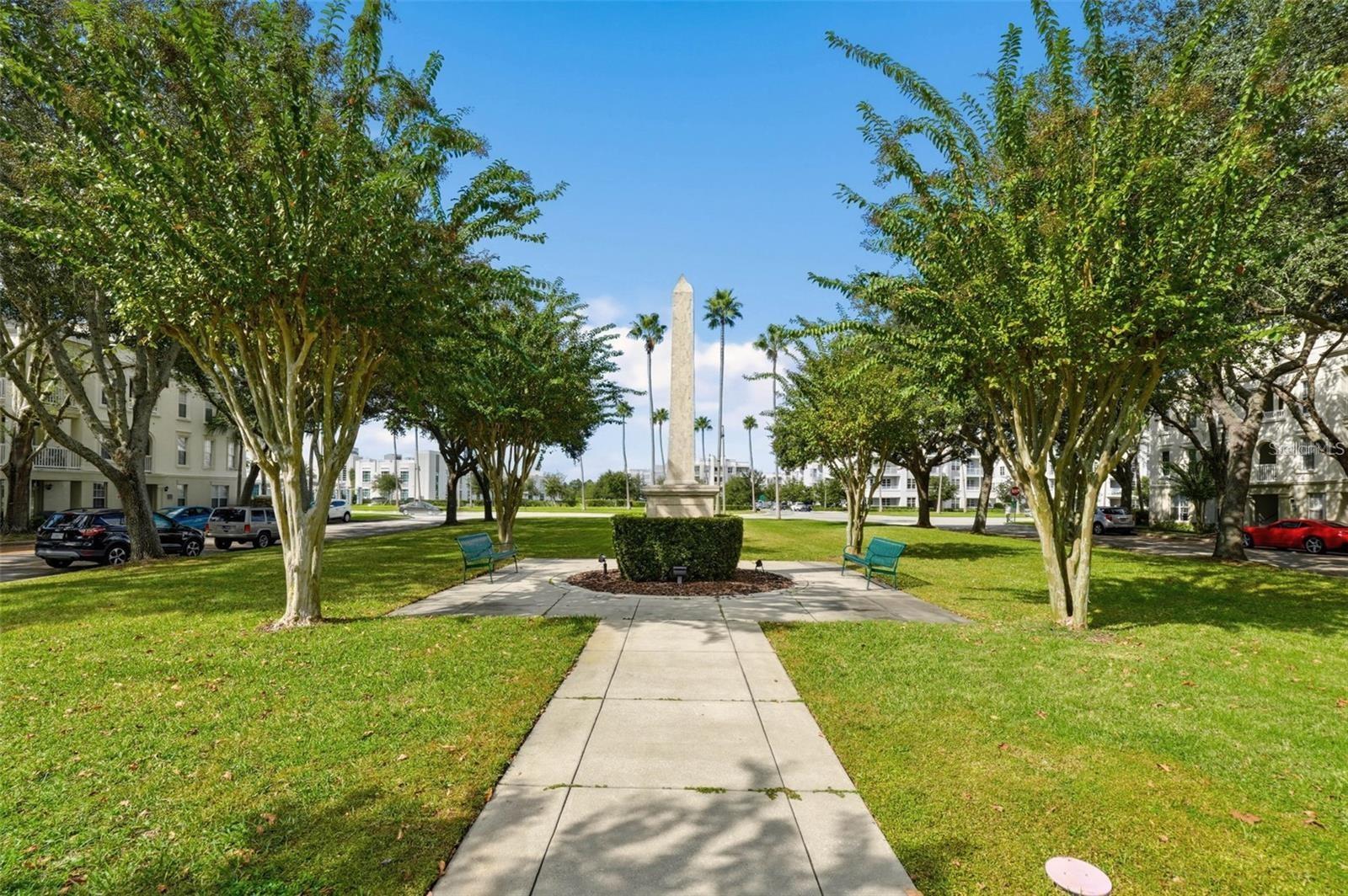 Residential community courtyard with a walkway, central stone monument, shade trees, and benches.