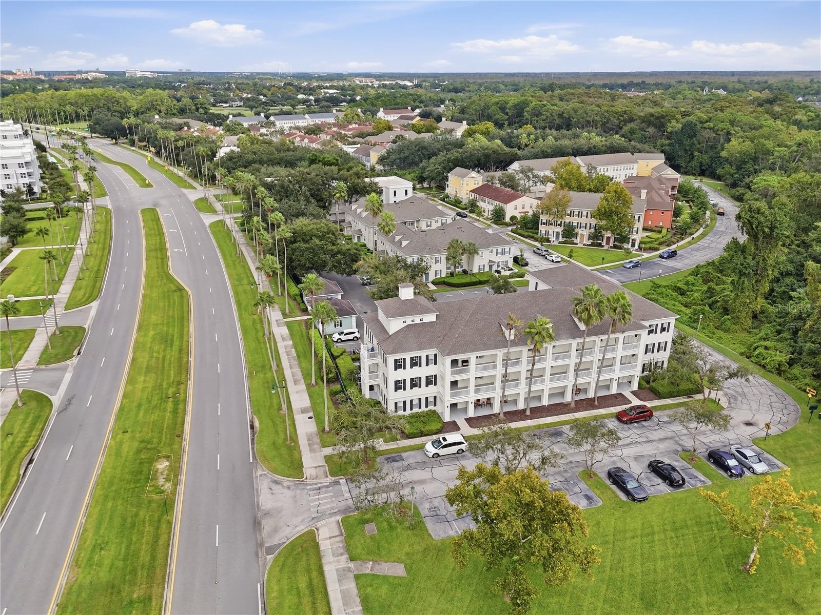 Aerial view of a white condominium building with covered balconies beside a palm-lined divided road.