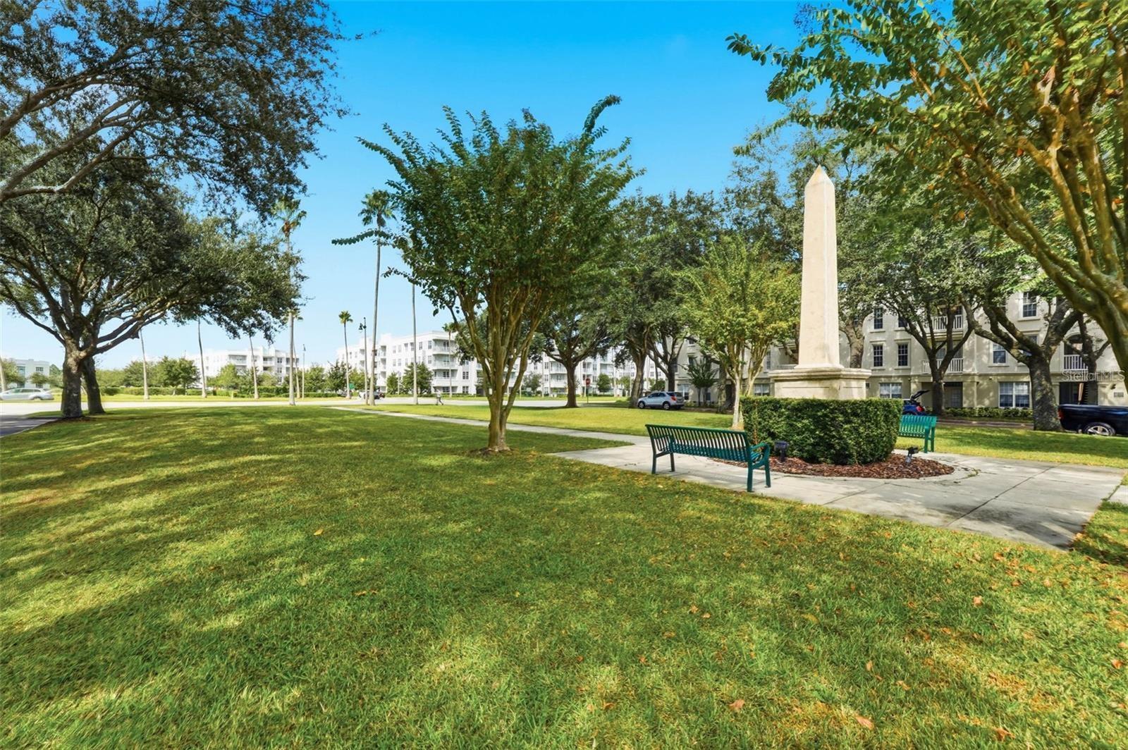 Landscaped community green with mature trees, benches, and a tall stone obelisk beside residential buildings.