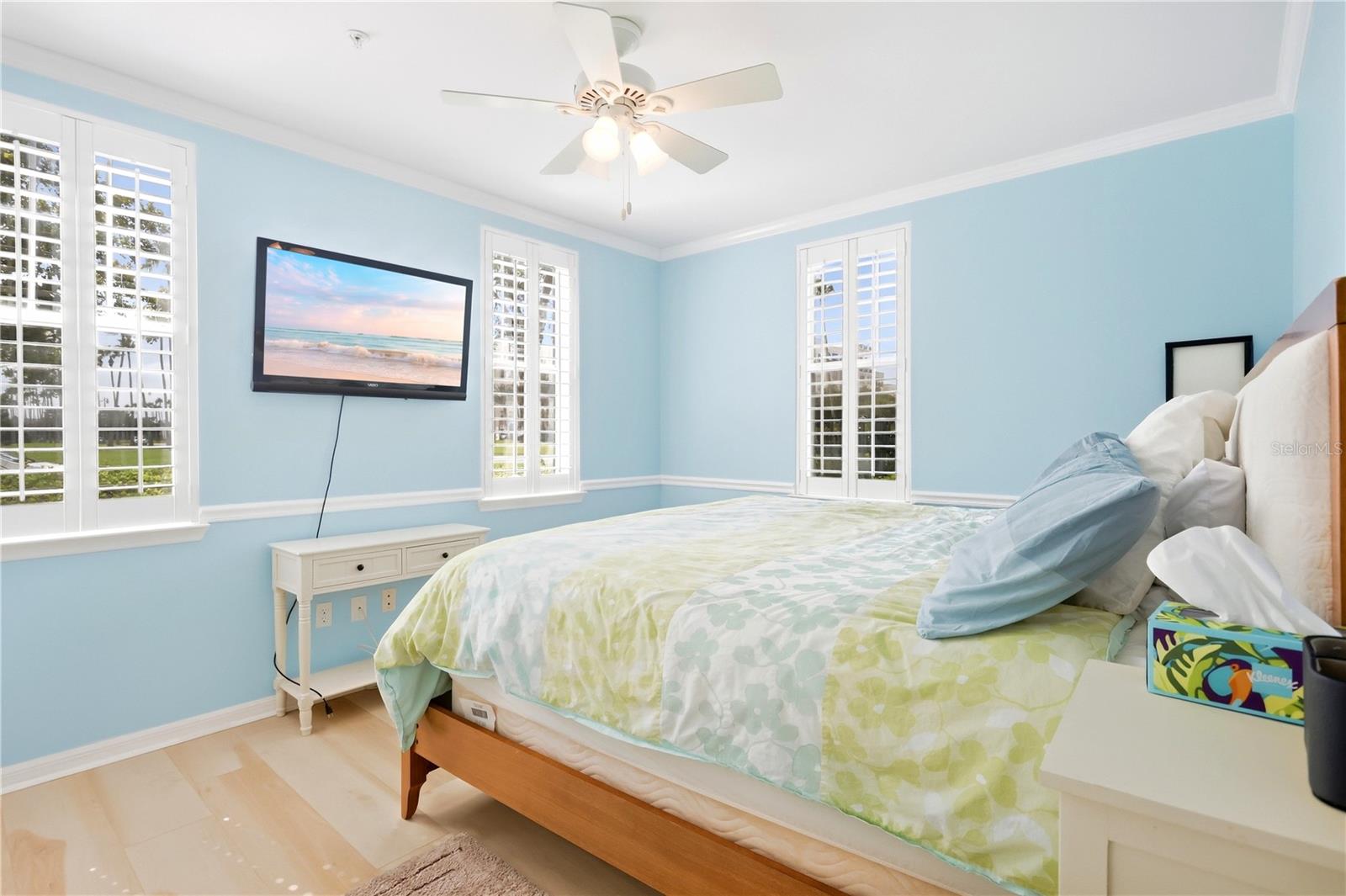 Bedroom with pale green bedding, light blue walls, plantation shutters, and a wall-mounted television.