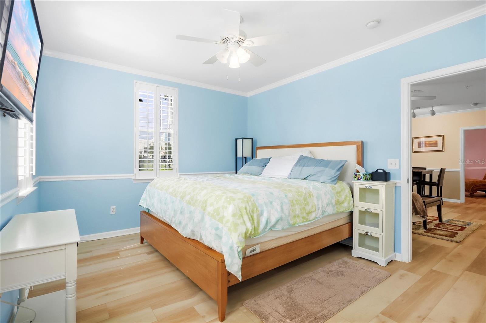 Bedroom with light blue walls, a wood-framed bed, shuttered windows, and an open doorway to the dining area.