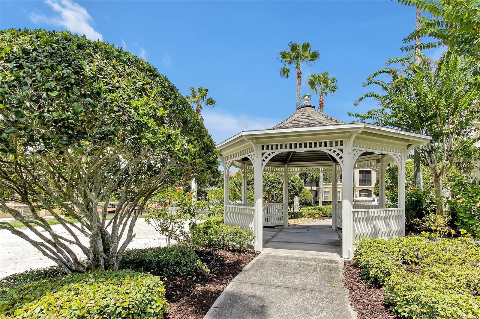 White decorative gazebo beside a landscaped walkway, with palm trees and residential buildings behind it.