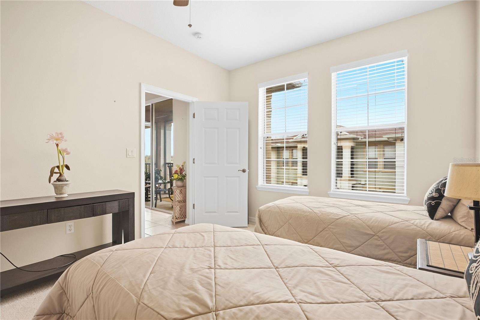 Bedroom with two beige quilted beds, a flower-topped console, and windows overlooking a neighboring building.