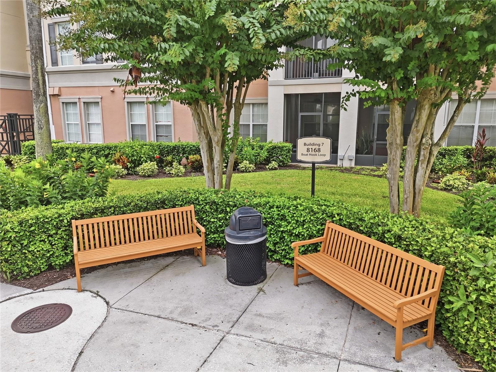 Condominium courtyard with two wooden benches, trimmed hedges, shade trees, and a Building 7 sign.