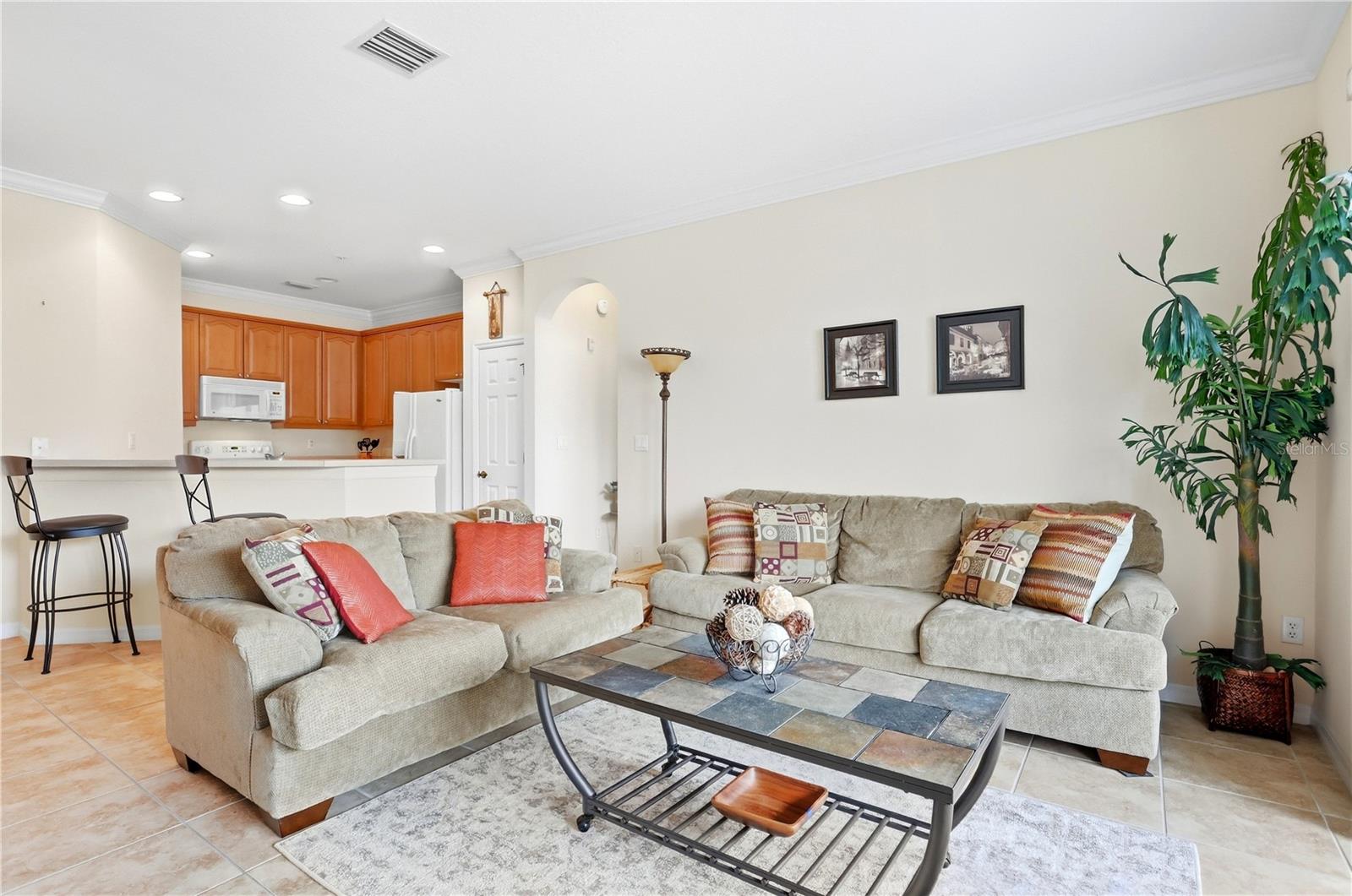 Living room with beige sectional sofas, a tiled coffee table, and an open kitchen with wood cabinets.