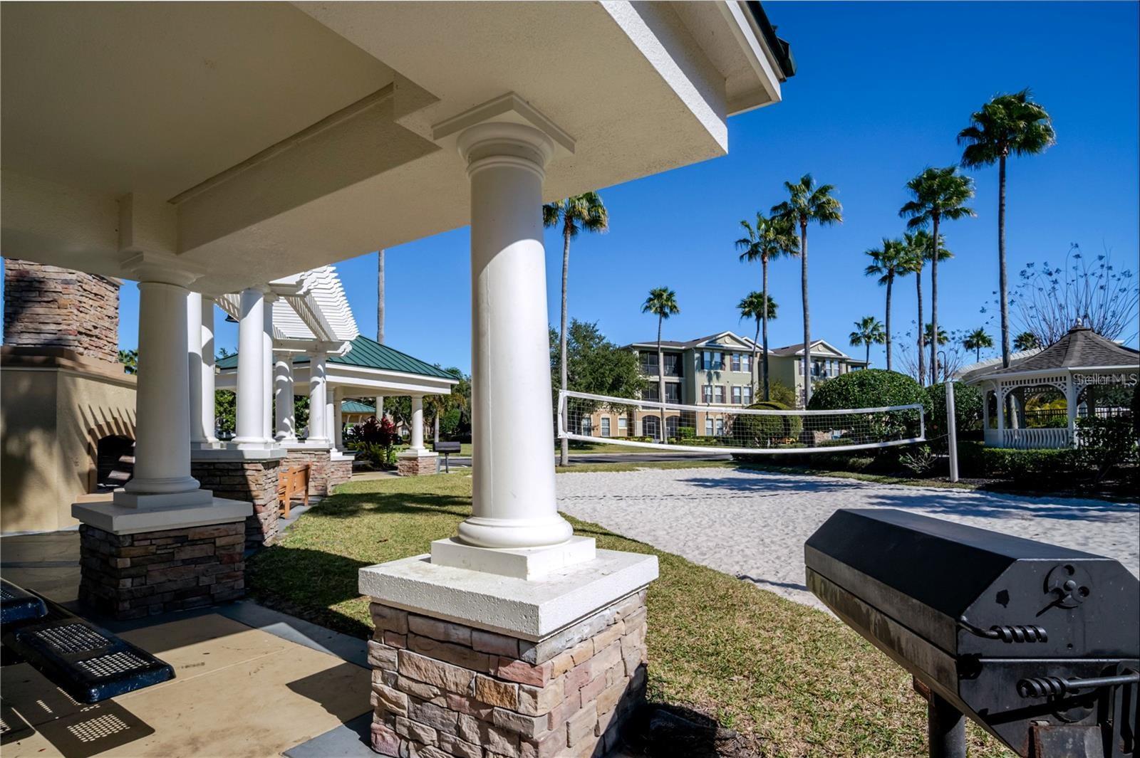 Community sand volleyball court beside a white-columned pavilion, gazebo, and palm trees.