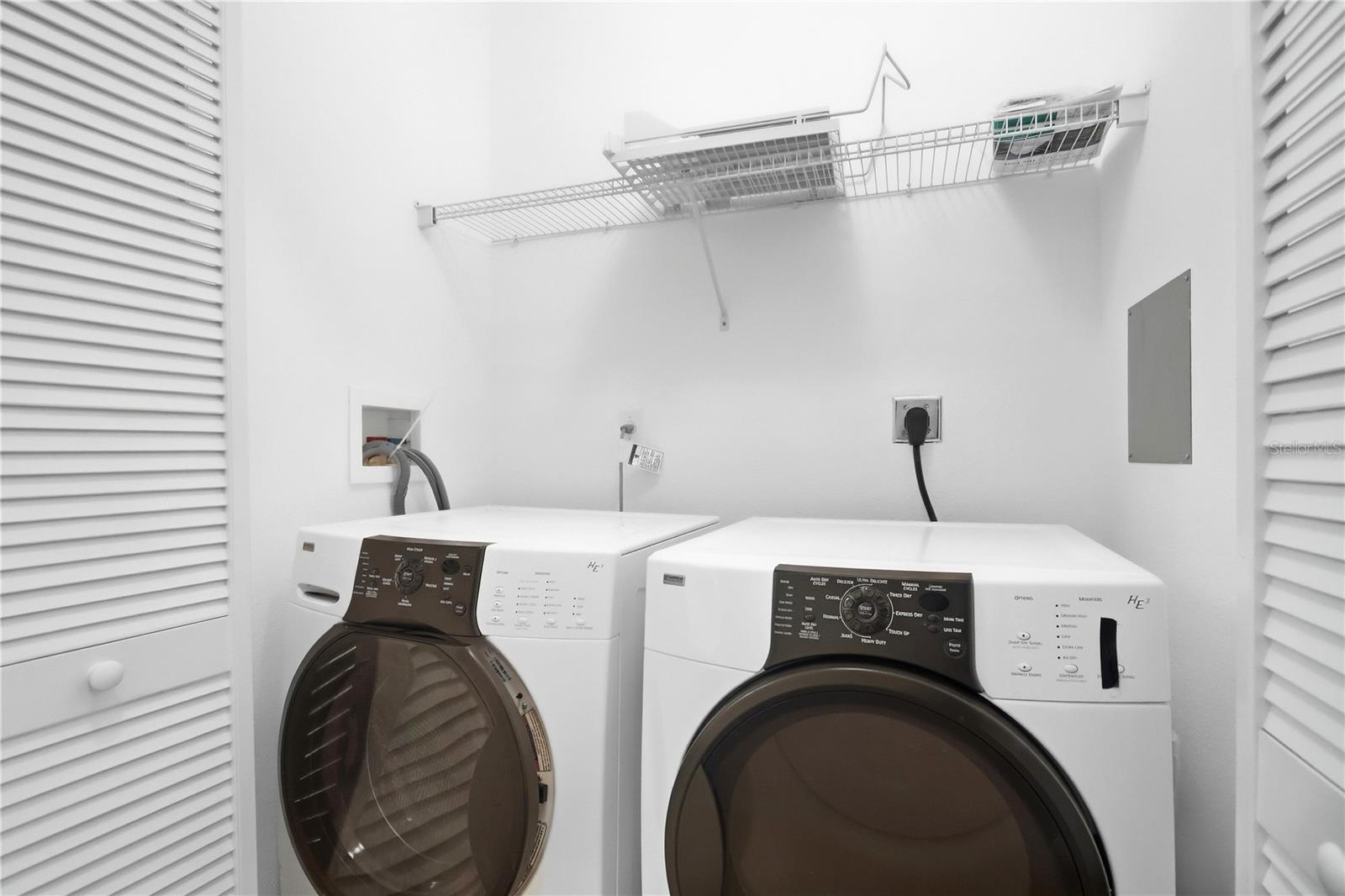 Laundry room with front-loading washer and dryer beneath a wire shelf, framed by white louvered doors.