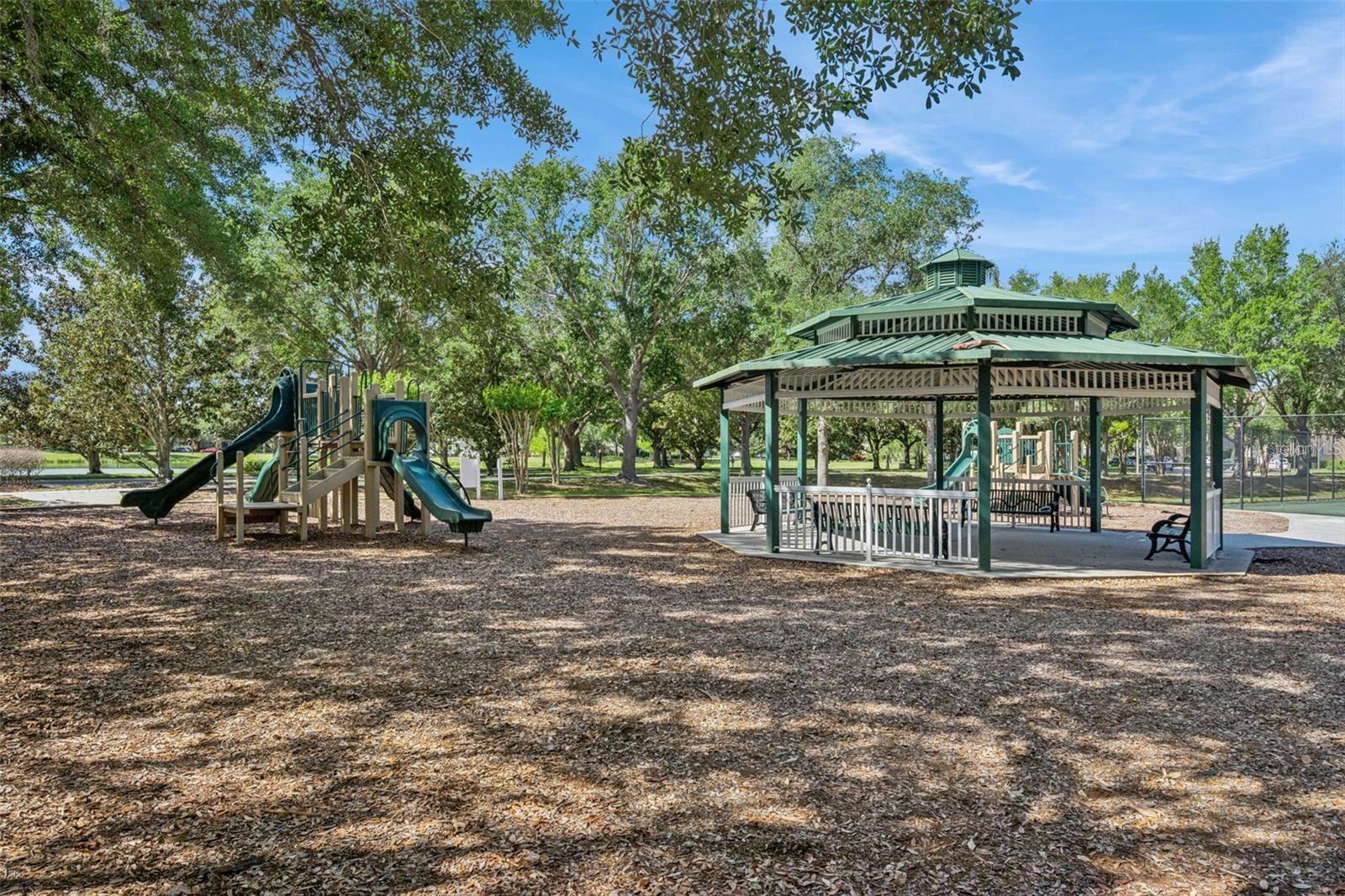 Community playground with a covered gazebo, benches, and a slide structure beneath mature trees.