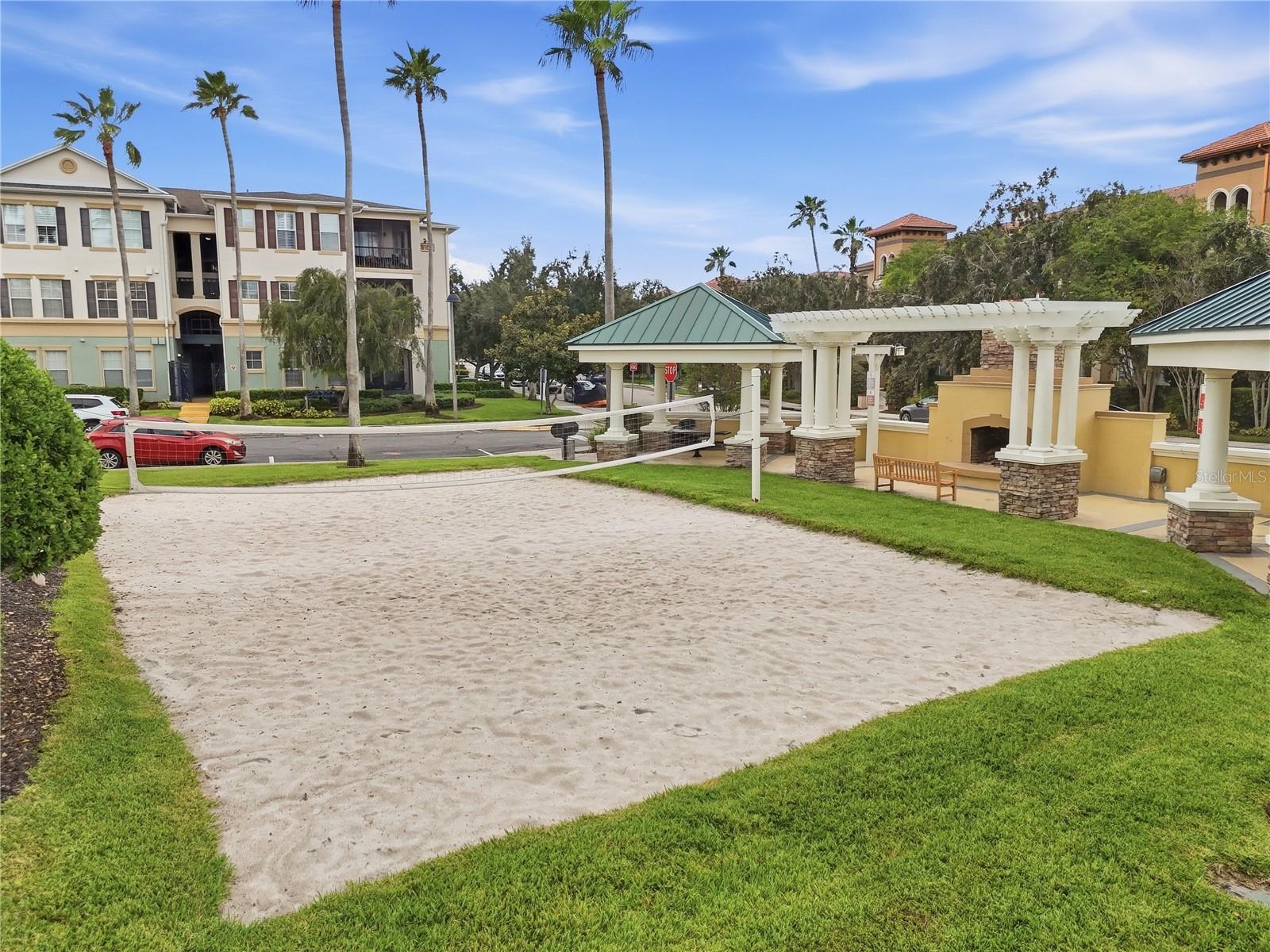 Community sand volleyball court beside a covered pavilion and palm-lined condominium buildings.
