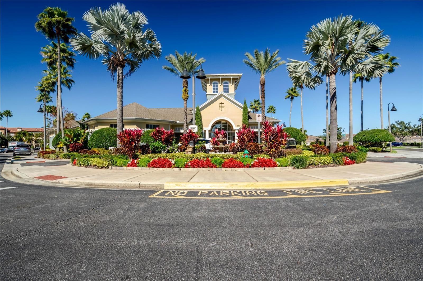 Clubhouse entrance framed by palm trees and colorful landscaping, with a no-parking marking on the drive.