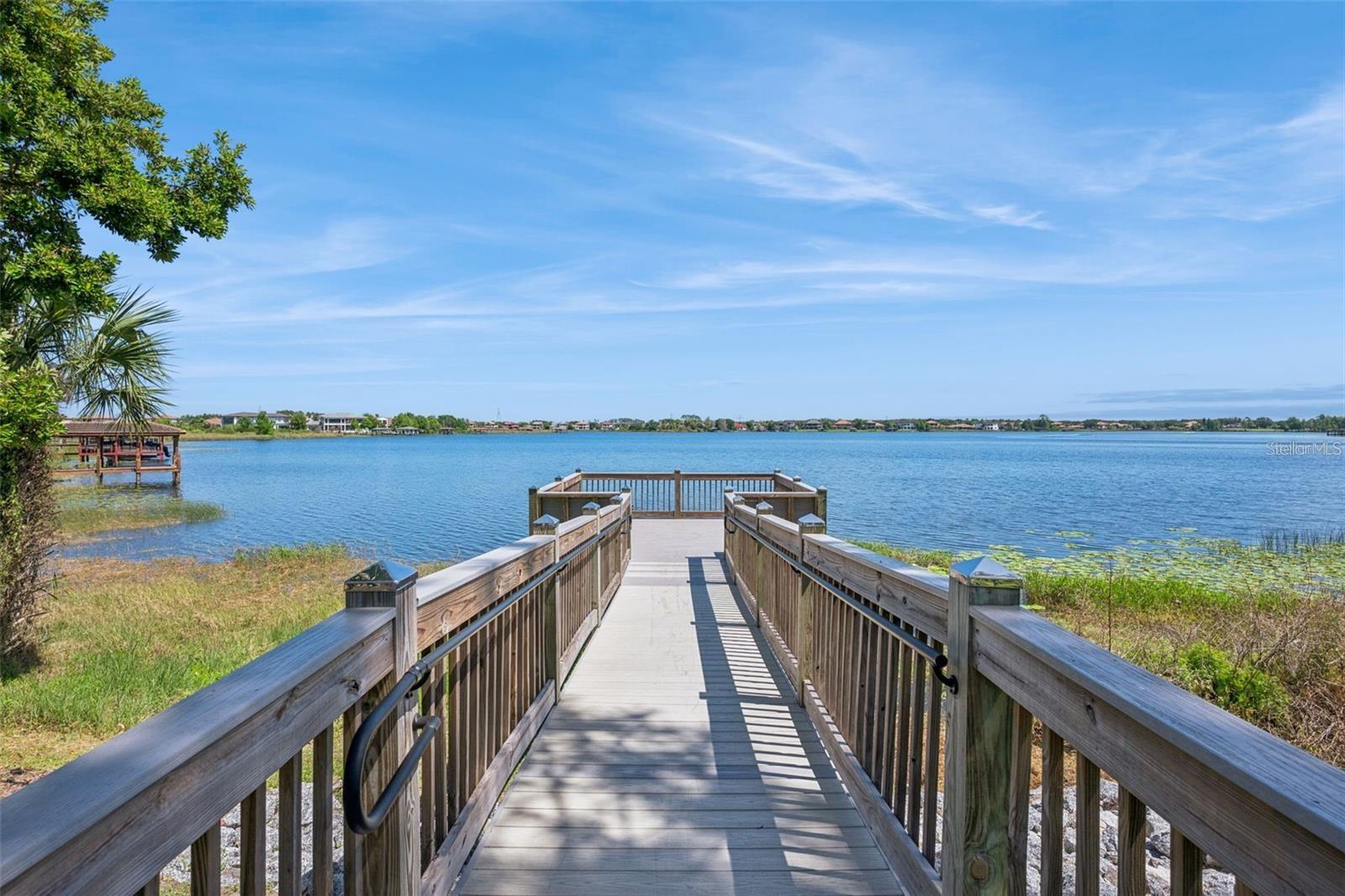 Wooden boardwalk extends to a lake, with shoreline homes visible across the water.