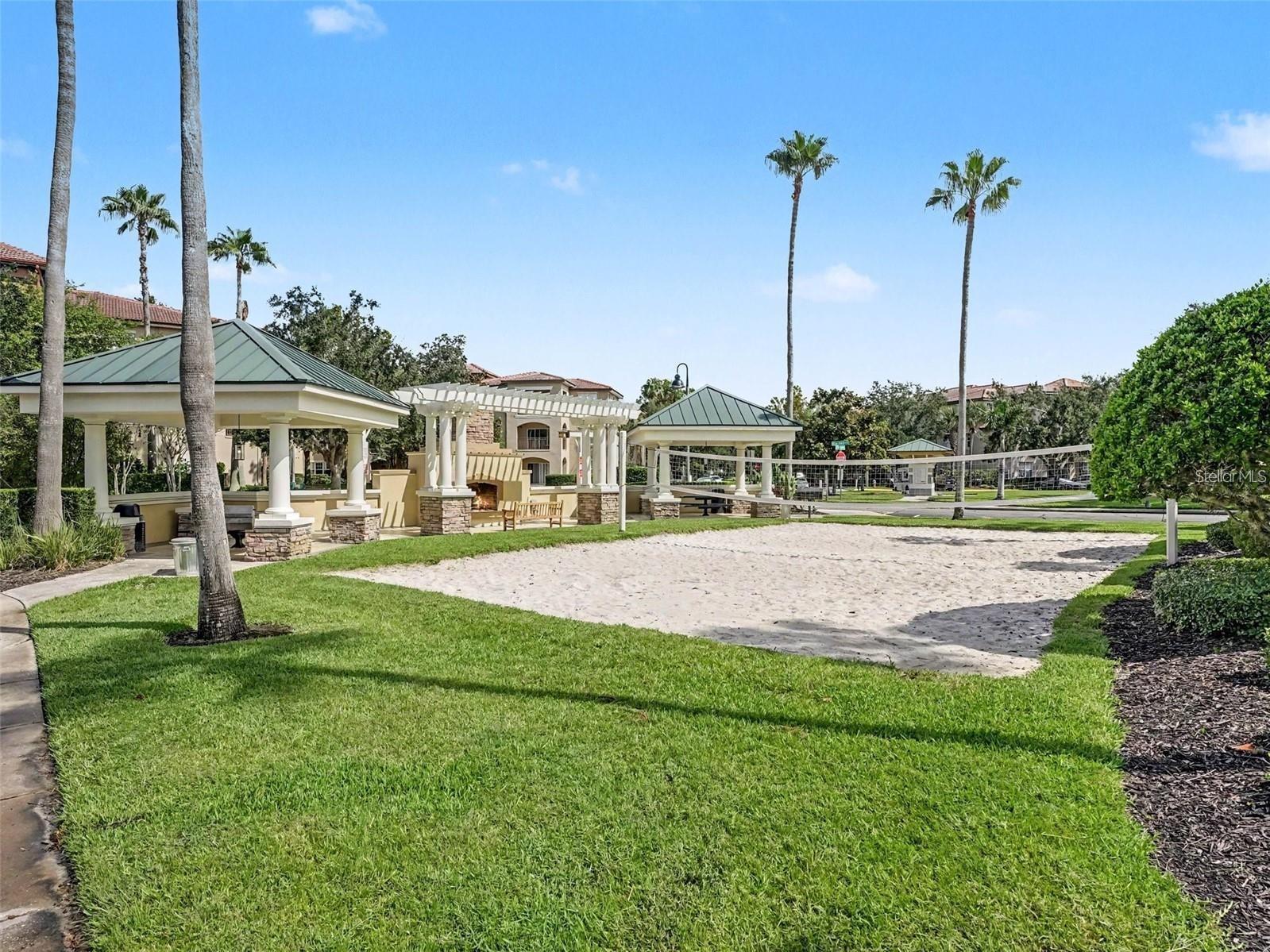Community sand volleyball court beside covered seating pavilions and palm trees.