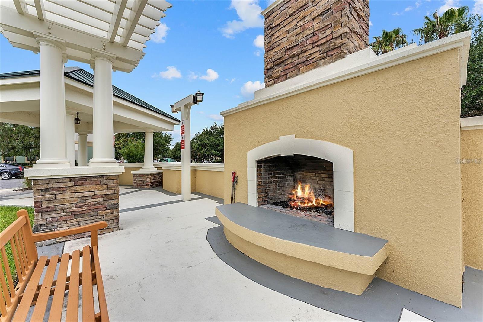 Outdoor patio with a burning stone fireplace, white-columned pergola, and wooden benches.