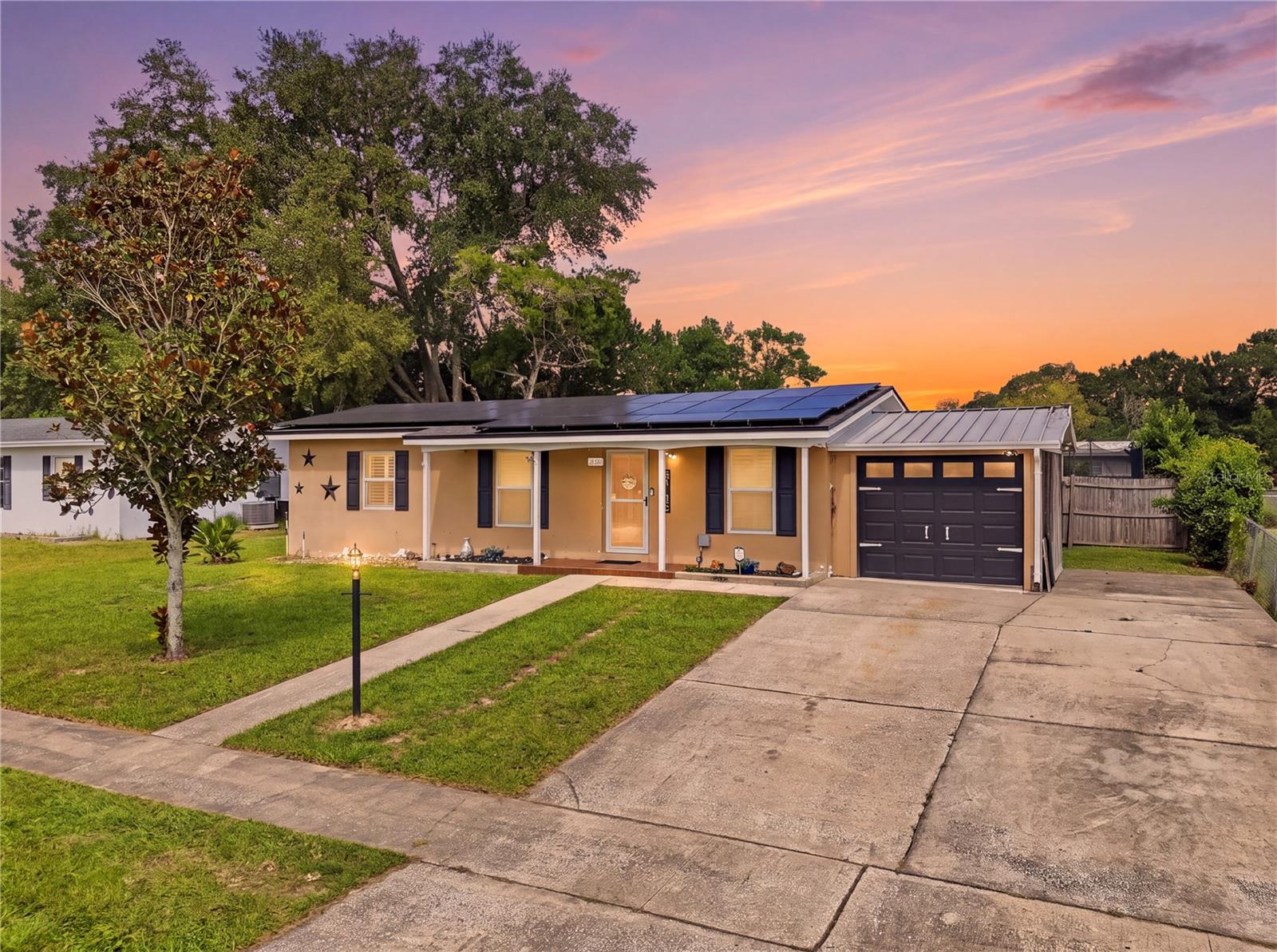 Single-story tan home with rooftop solar panels, a dark garage door, and a broad driveway at dusk.