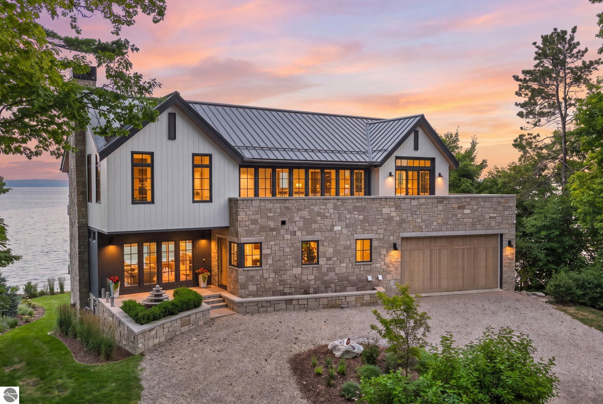 Modern stone-and-siding lakeside home at dusk, with warm glowing windows and a metal roof.