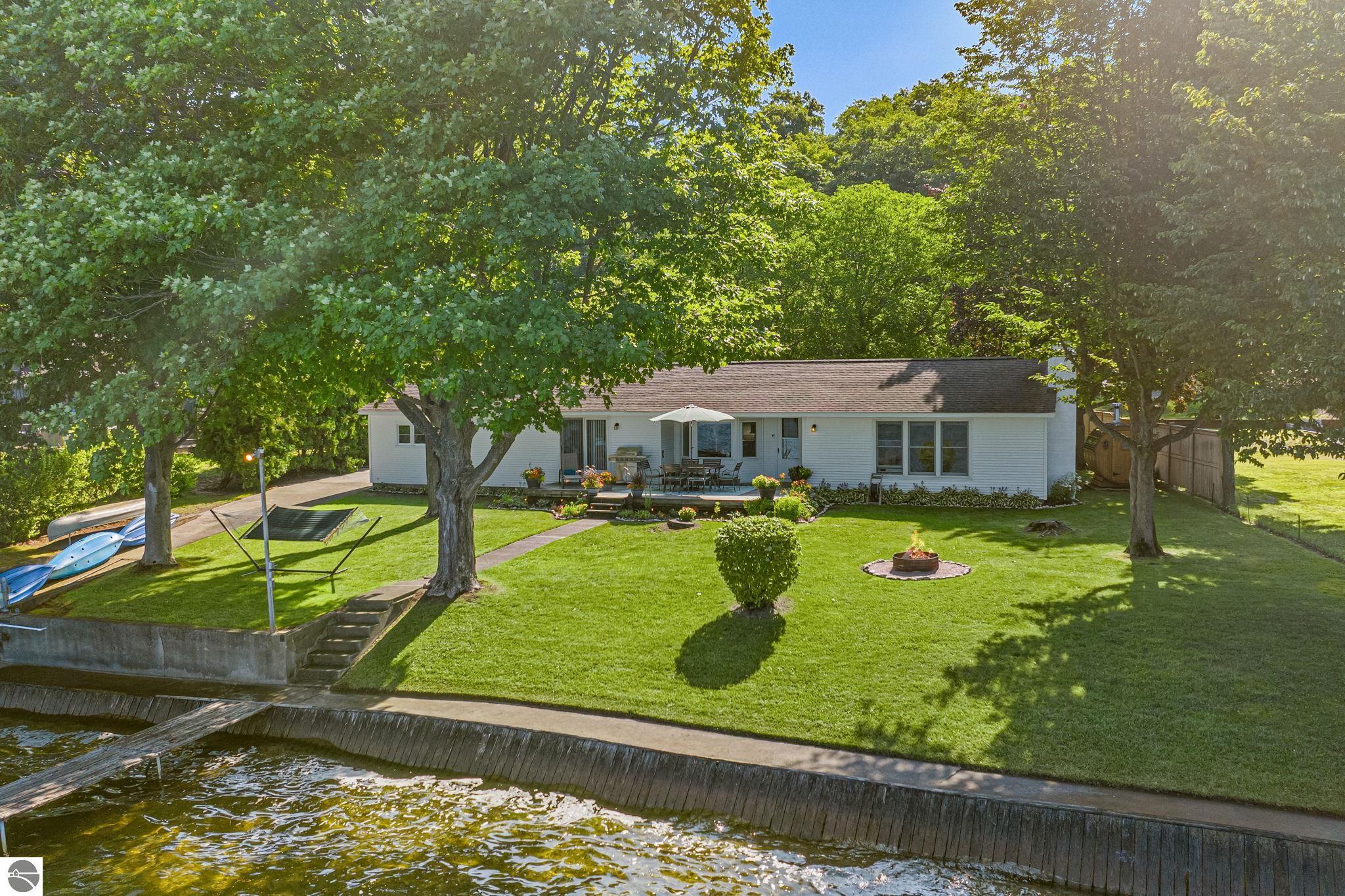 Waterfront home with a broad tree-shaded lawn, outdoor dining area, fire pit, and kayaks by the shore.