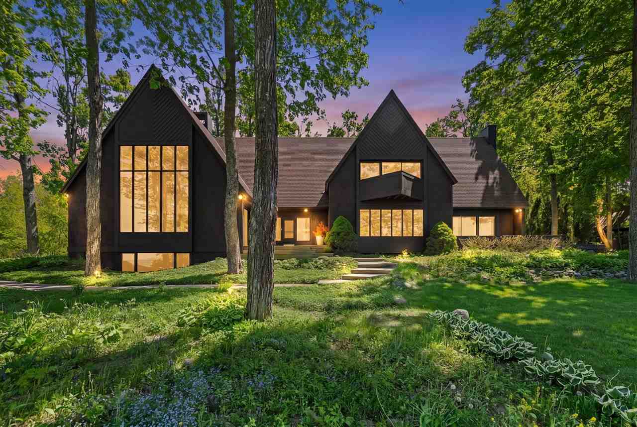 Modern dark-gabled house with glowing windows among mature trees and landscaped gardens at dusk.