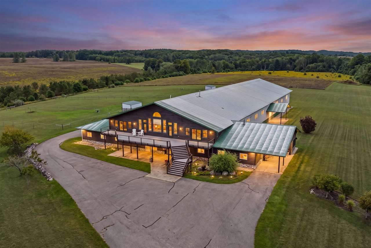 Large country home with a raised deck and curved driveway, surrounded by fields at dusk.