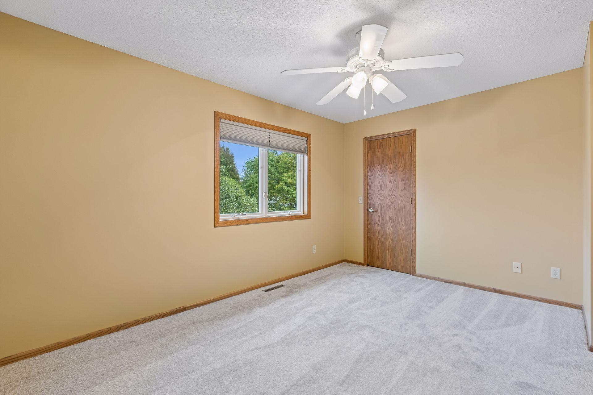 Empty bedroom with beige walls, gray carpet, a tree-view window, and a white ceiling fan.