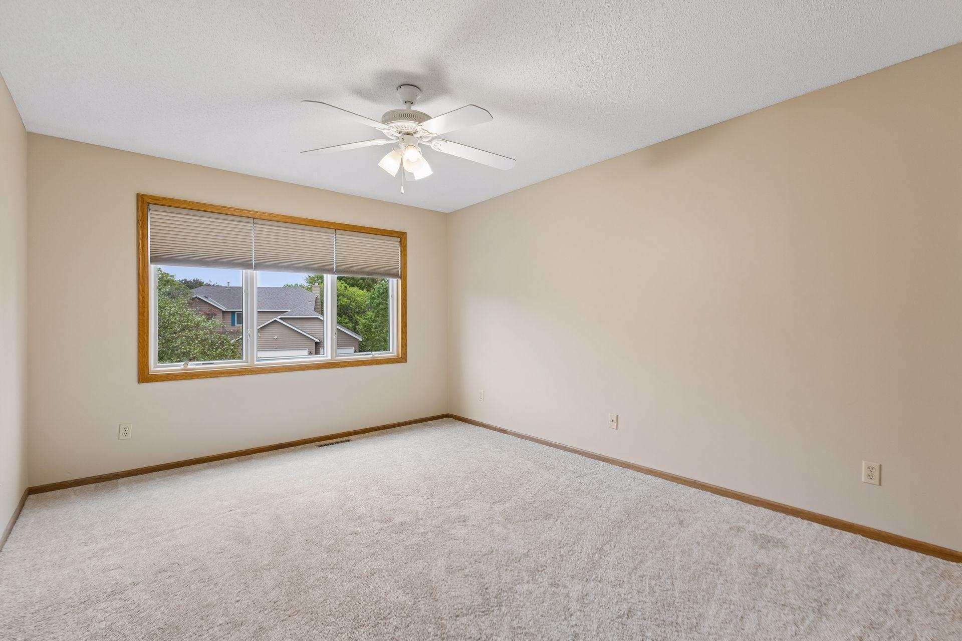 Empty carpeted bedroom with a wide window, blinds, and a white ceiling fan.