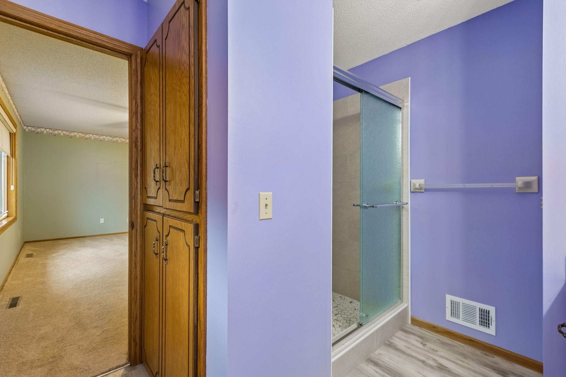 Bathroom with purple walls, glass shower, and built-in wooden cabinet beside an adjoining room.