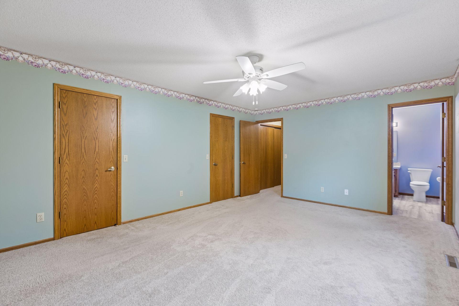 Empty carpeted bedroom with pale blue walls, a ceiling fan, and an adjoining bathroom.