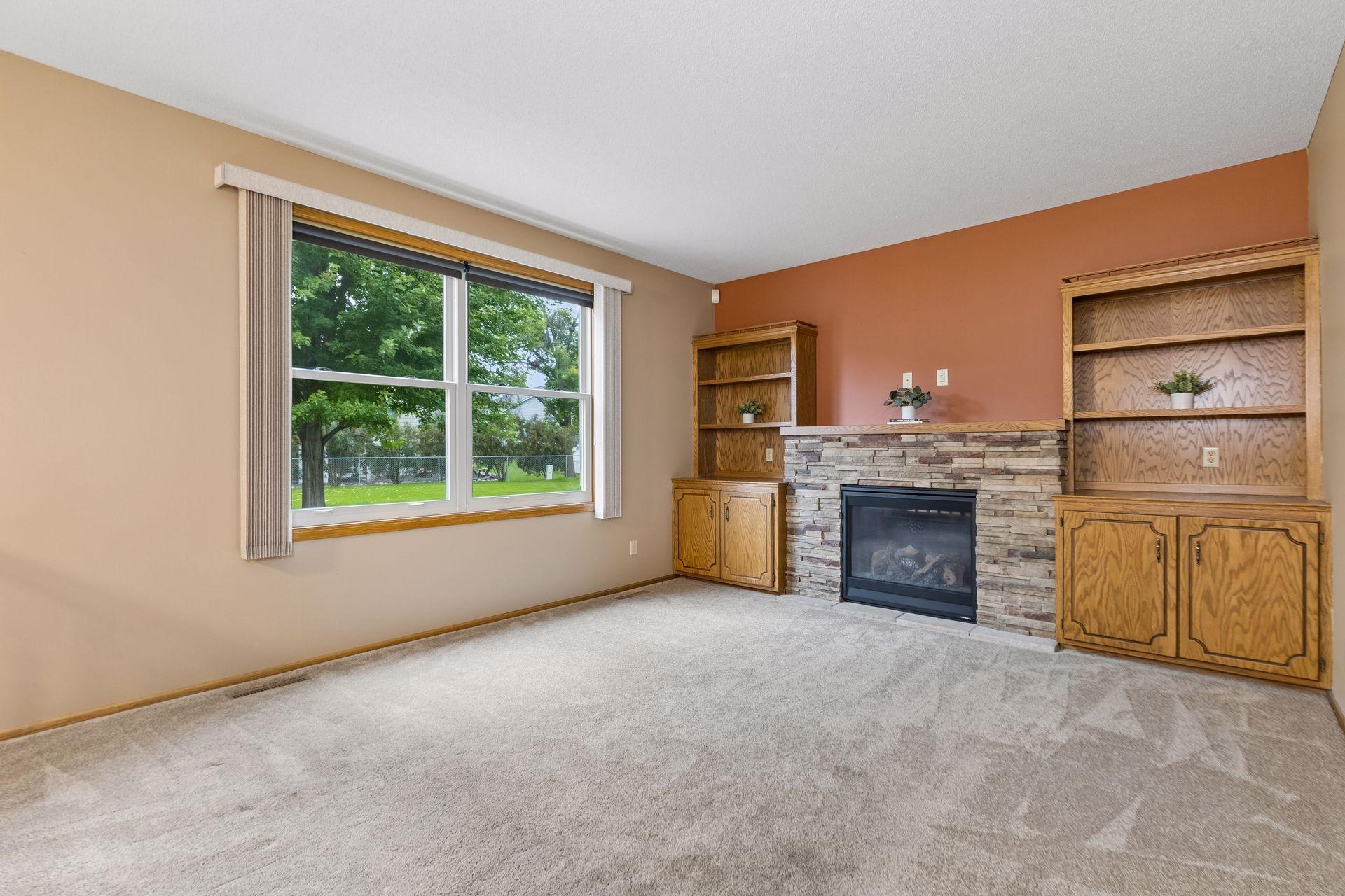 Living room with a stone fireplace, built-in oak shelving, and a large window overlooking a green yard.