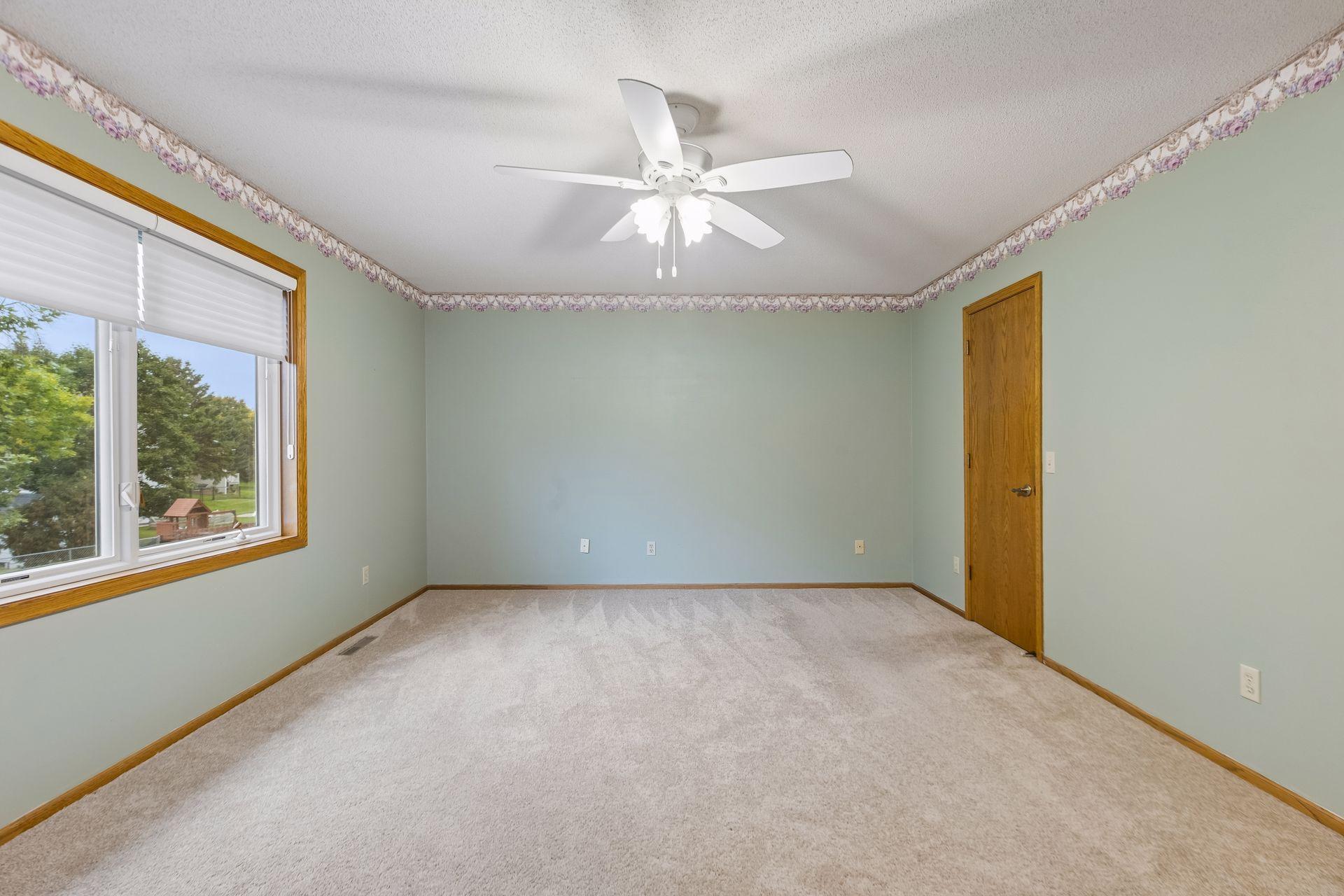 Empty bedroom with pale green walls, light carpet, a wood-trimmed window, and a white ceiling fan.