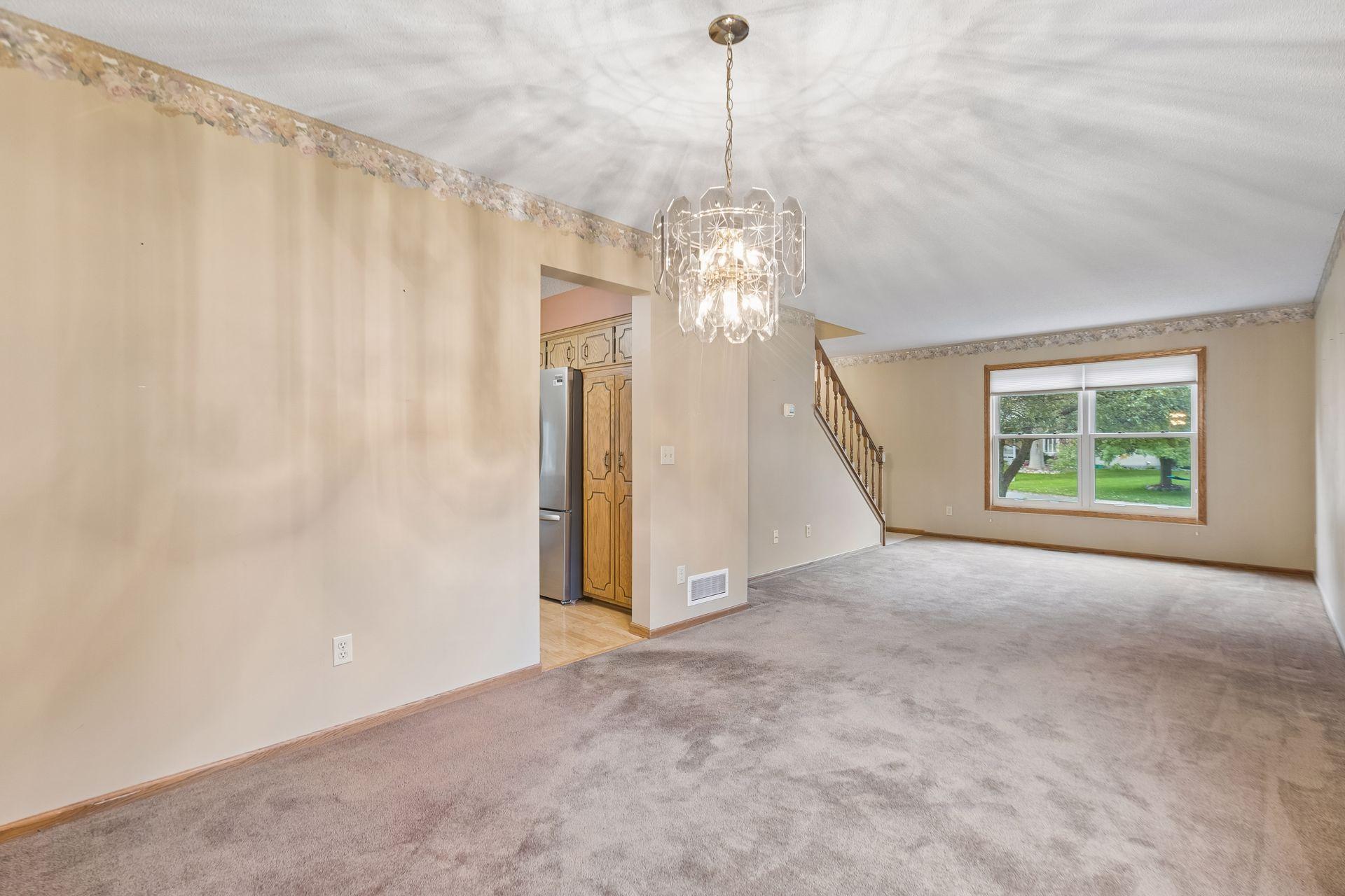 Empty carpeted living room with a chandelier, staircase, and large window.