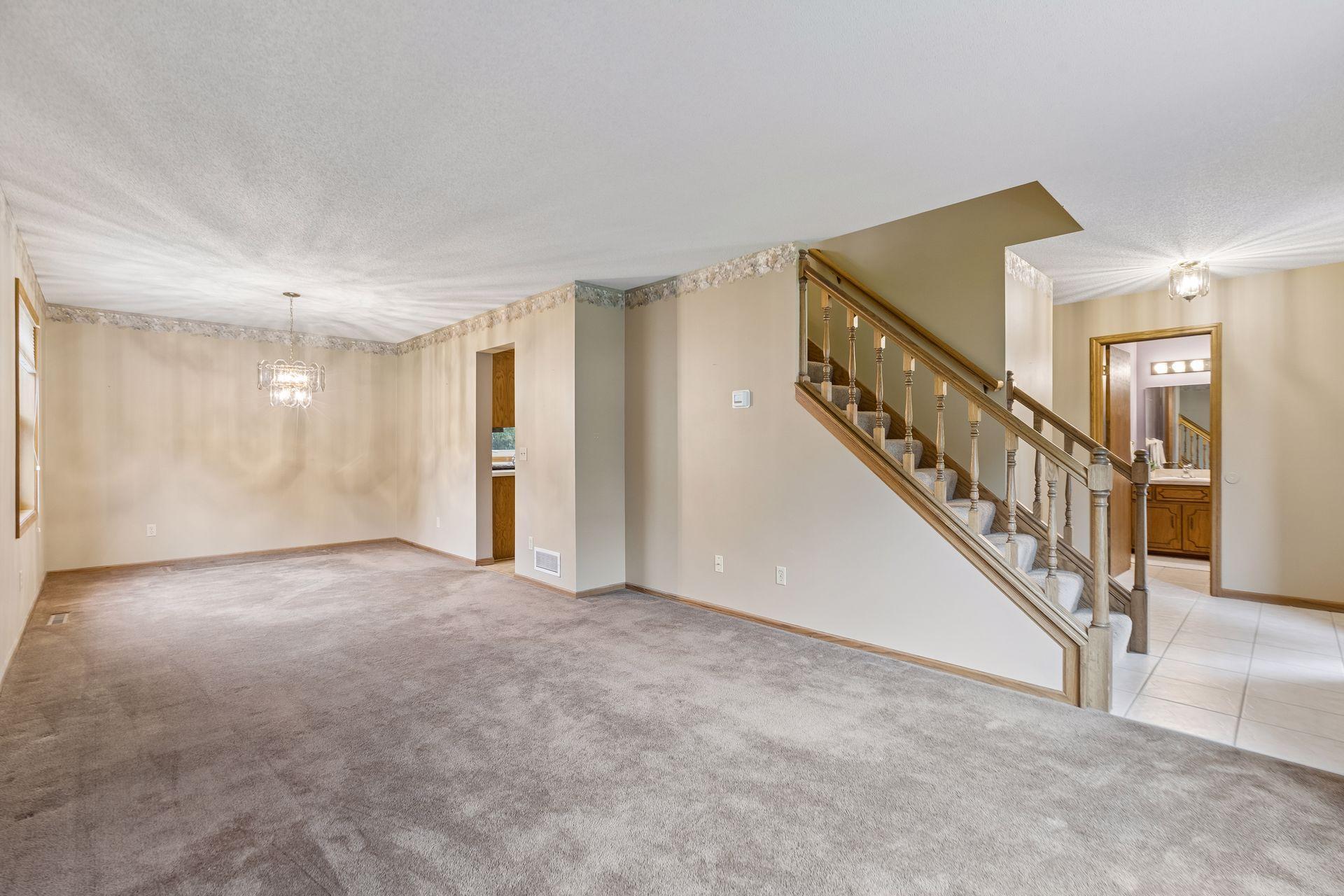 Vacant carpeted living and dining area with a chandelier and wooden staircase.