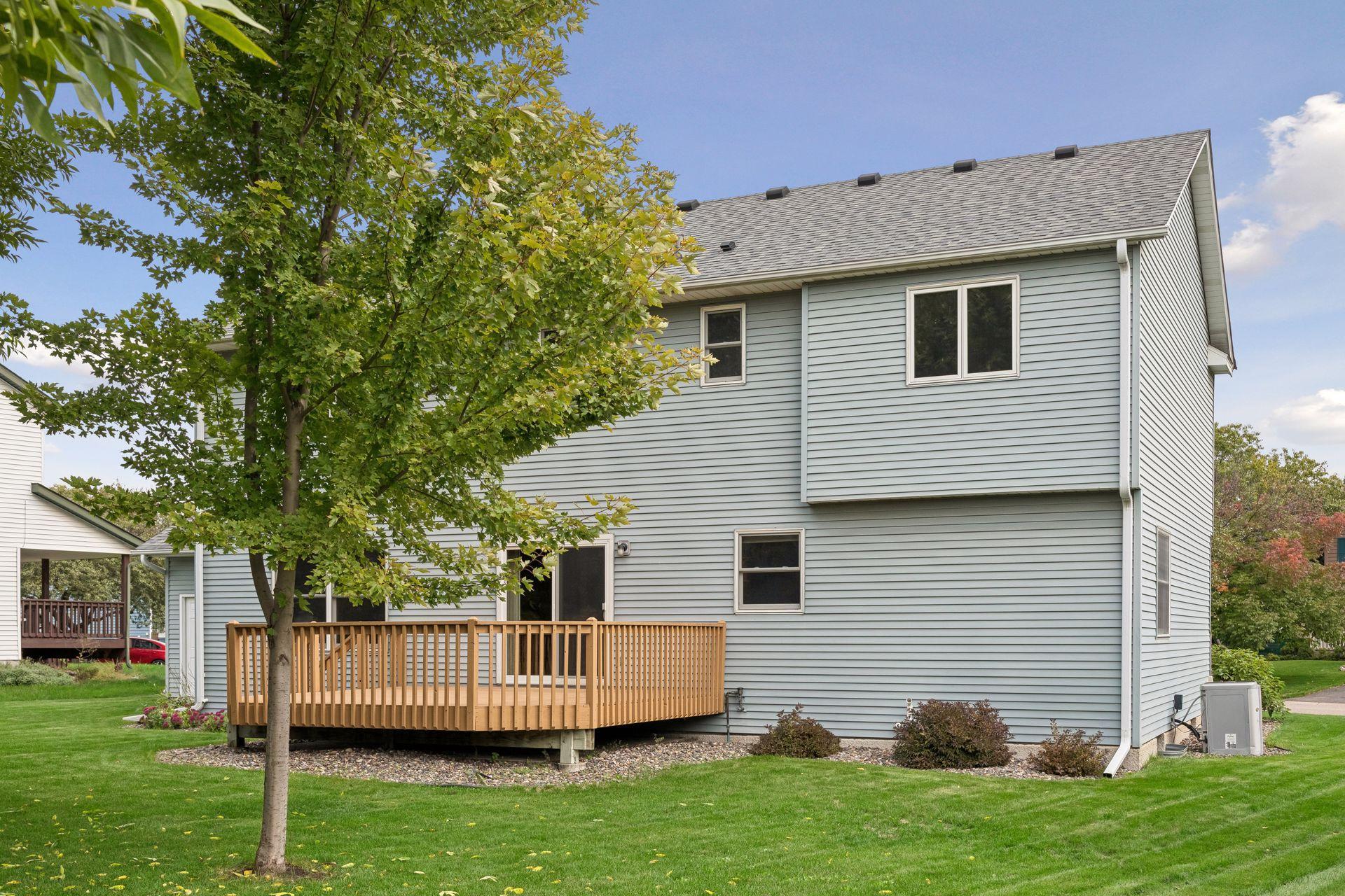 Two-story sided home with a wooden deck, partly screened by a leafy tree.