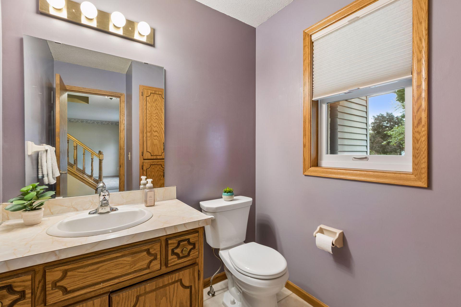 Bathroom with a wood vanity, large mirror, mauve walls, and a window with a partly lowered blind.