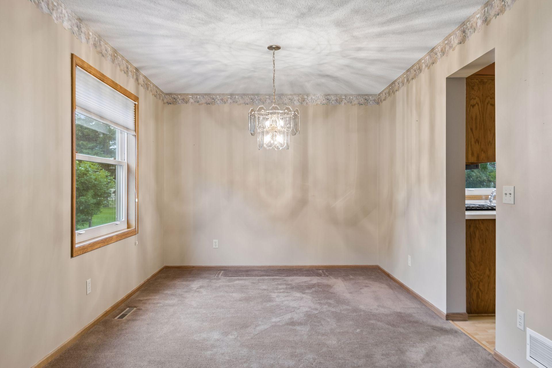 Empty dining room with a crystal chandelier, gray carpet, and a wood-trimmed window.
