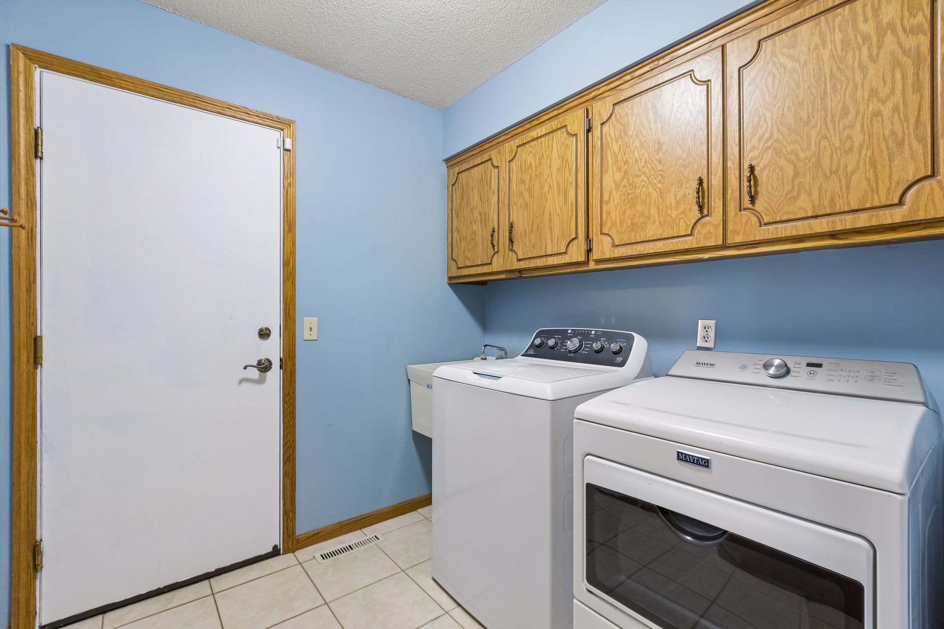 Laundry room with washer and Maytag dryer beneath oak cabinets, beside a white door.