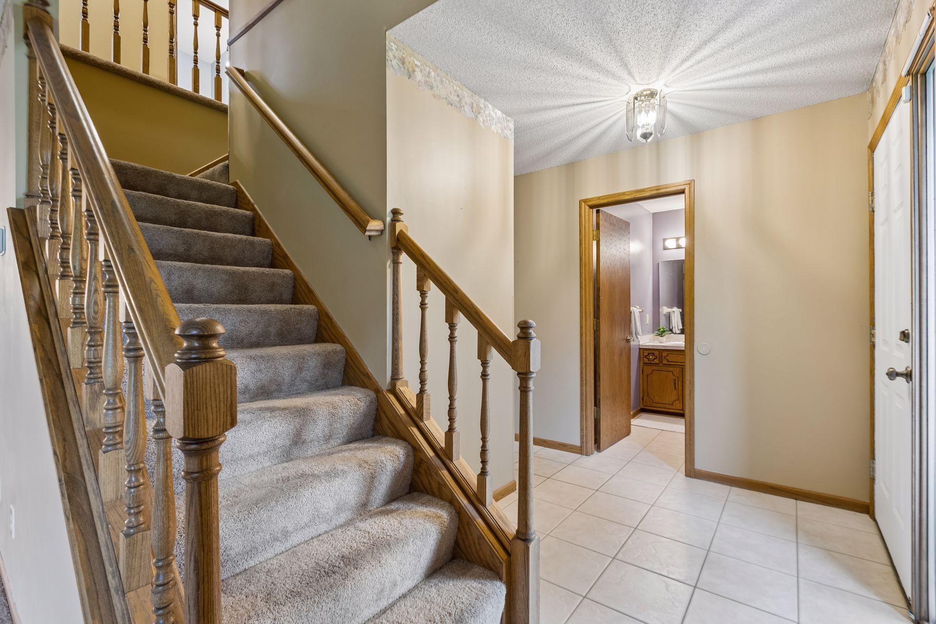 Entry hall with a carpeted staircase, wooden railings, and tiled floor beside an open bathroom doorway.