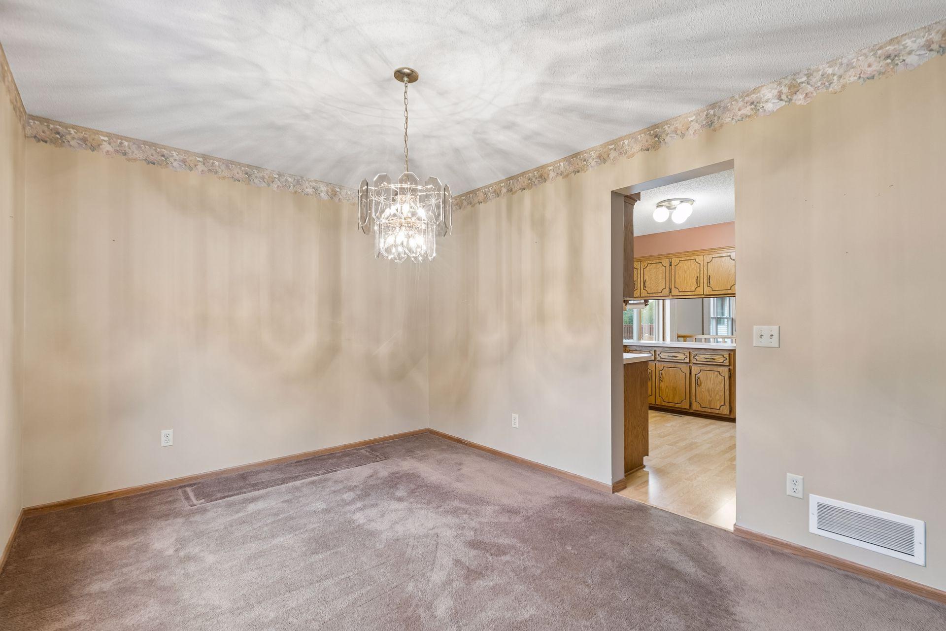 Empty carpeted dining room with a crystal chandelier and doorway to a wood-cabinet kitchen.
