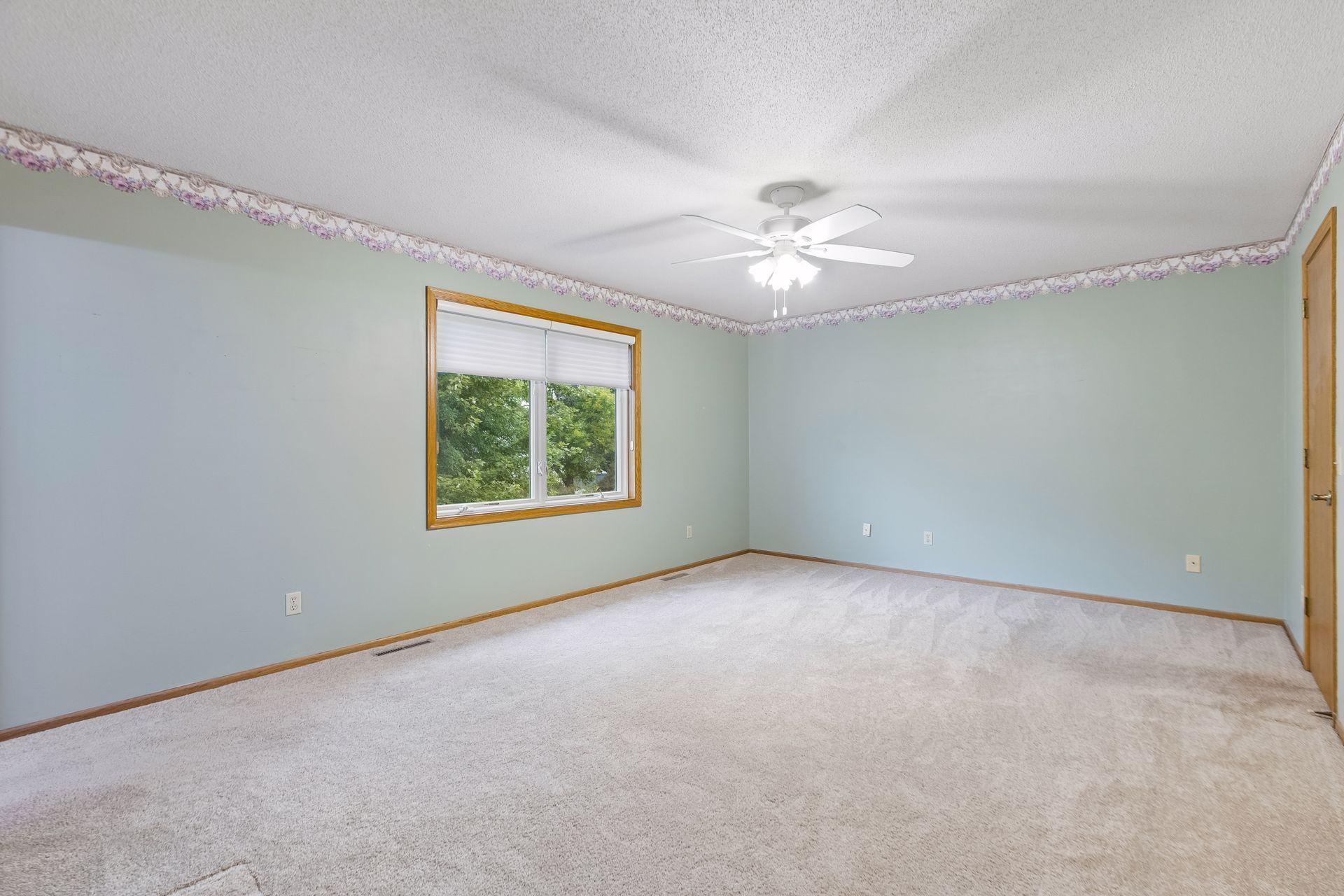 Empty carpeted bedroom with pale green walls, a window with blinds, and a ceiling fan.