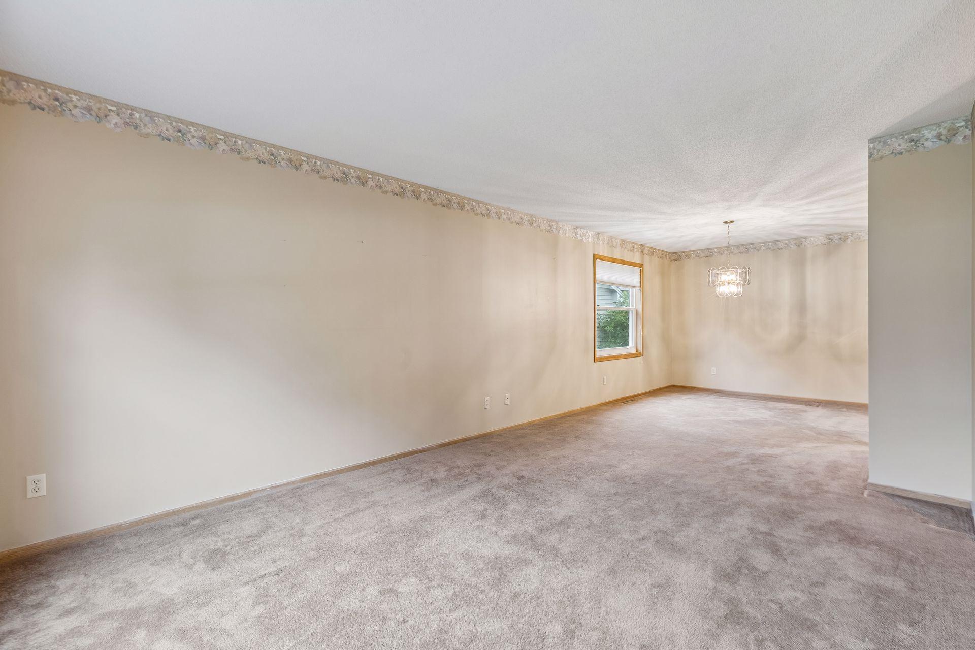 Empty carpeted living and dining room with a wood-trimmed window and chandelier.